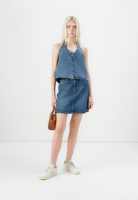 Farmer nadrág szett, amely egy gombos, rövidített pántos felsőből és egy testhezálló szoknyából áll. Világoskék szín, sima textura, fehér sneaker cipőkkel és barna táskával párosítva.