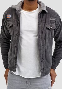 Grijze corduroy jas met een kraag gevoerd met sherpa, voorzien van knoopsluitingen en twee voorzakken. Draagt over een wit T-shirt.