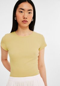 Tæt siddende kortærmet t-shirt i lys gul ribbet stof, rund halsudskæring, glat tekstur, båret med en hvid nederdel med elastisk talje.