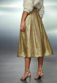 Frau in einem strukturierten goldenen Midirock, einem flauschigen cremefarbenen Pullover und metallisch goldenen High-Heels, die mit einer entspannten Hand steht.