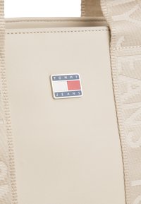 Beige tote-taske med struktureret side og Tommy Jeans-logo i rød, hvid og blå. Den har robuste stropper og en glat finish.