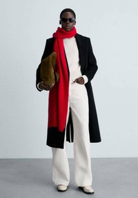 Zwarte jas, rode sjaal, crèmekleurige trui en wijde witte broek. Houdt bruine schoenen vast. Zonnebril en een strakke haarstijl maken de look compleet.