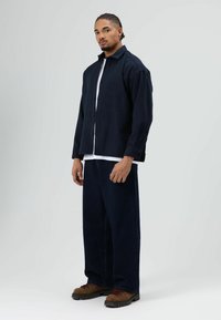 Marineblauw overhemdd met knopen van gestructureerde stof, gedragen over een wit T-shirt. Donkerblauwe spijkerbroek en bruine laarzen maken de outfit compleet.