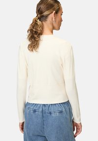 Helles beige Langarmoberteil mit strukturierten Ärmeln und taillierter Passform, kombiniert mit lockeren blauen Jeanshose mit elastischem Bund und Taschen.