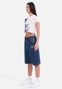 Camiseta rosa recortada con diseño gráfico, combinada con pantalones cortos de mezclilla azul bermuda que tienen detalles coloridos en los bolsillos y zapatillas blancas.