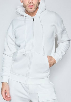 Homme portant un sweat à capuche blanc zippé avec cordons et pantalon blanc assorti, une main dans la poche du sweat, sur un fond clair uni.