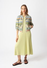 Mezgimo bomberis su sudėtingais daugiataškių raštais, dėvimas virš šviesiai žaliai raukšlėtos suknelės. Rudi sandalai su dirželiais puikiai užbaigia įvaizdį.