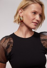 Black ribbed top with sheer black puff sleeves, paired with large reflective gold earrings, worn by a person with short blonde hair.