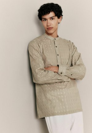 Hellbeiges Hemd mit Stehkragen und dezentem Muster. Lange Ärmel und Knopfleiste, aus einem strukturierten Stoff gefertigt.