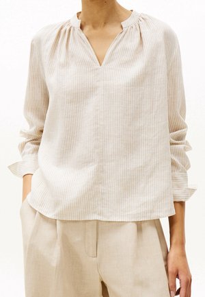 Vrouw die een beige gestreepte blouse met lange mouwen, V-hals en opgerolde mouwen draagt, gecombineerd met beige broek, handen in de zakken.