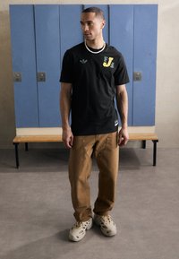 Black t-shirt with green and yellow graphic logo; brown loose-fit pants; light cream footwear; plain design; indoor setting with lockers.