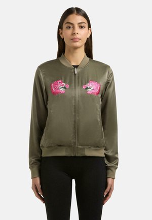 Kobieta ubrana w oliwkowo-zieloną kurtkę bomber z różowymi, haftowanymi głowami tygrysów na piersi, w połączeniu z czarnymi spodniami, stojąca na białym tle.