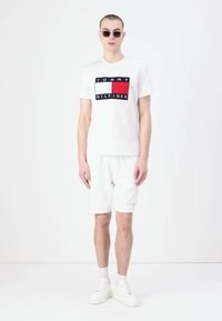 Balta medvilninė marškinėliai su raudonu, baltu ir mėlynu logotipu; baltos šortai; baltos sportbačiai; apvalūs saulės akiniai; modelis stovi prieš vienodą foną.