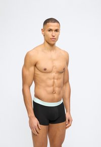 Μυώδης άνδρας με κοντά μαλλιά, φορώντας μαύρα ανδρικά εσώρουχα τύπου μποξερ με ανοιχτόχρωμη ζώνη μέσης, στέκεται μπροστά σε απλό λευκό φόντο.