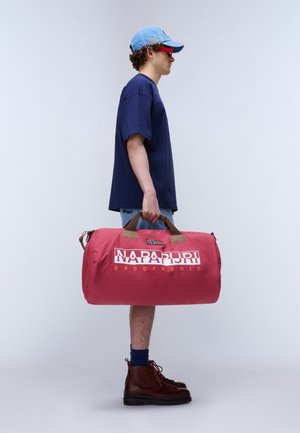 Jeune personne portant une casquette bleue, une chemise marine, un short en denim et des bottes, tenant à la main un grand sac de sport rouge Napapijri.