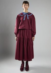 Abito bordeaux con maniche lunghe e gonna arricciata. Presenta un colletto blu e un dettaglio a fiocco, realizzato in tessuto liscio, abbinato a scarpe coordinate.