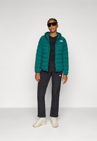 Teal dunjakke med hette, svart t-skjorte, svarte bukser og beige joggesko; har logodetaljer og en tettsittende design.