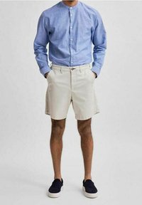 Camicia a strisce blu chiaro con bottoni, pantaloni corti beige e scarpe slip-on blu navy. La camicia ha un colletto a banda, mentre i pantaloni corti hanno tasche laterali.