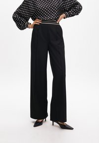 Black wide-leg trousers with sharp pleats, paired with a polka dot, puff-sleeved top featuring a striped elastic waistband and pointed black heels.