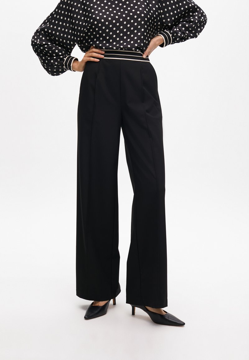 Black wide-leg trousers with sharp pleats, paired with a polka dot, puff-sleeved top featuring a striped elastic waistband and pointed black heels.