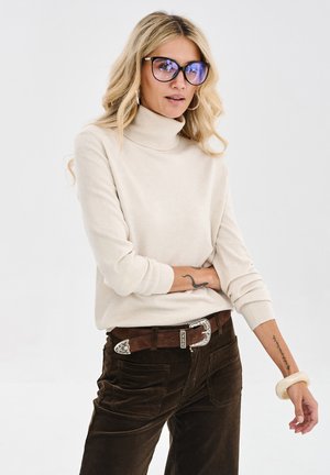 Kobieta z długimi blond włosami nosząca duże okulary z ciemnymi oprawkami, beżowy sweter z golfem, brązowe spodnie i pasek, pozująca na białym tle.