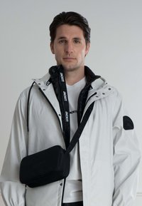 Bílá nepromokavá bunda, černá mikina na zip pod ní, černá taška přes rameno s embosovaným logem, nošena přes bílý tričko.