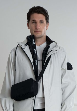 Hvid vandtæt jakke, sort hættetrøje med lynlås under, sort skuldertaske med præget logo, båret over en hvid T-shirt.