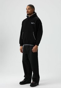 Zwarte hoodie met decoratieve stiksels, een kangaroezak en een losse pasvorm. Gecombineerd met loszittende zwarte jeans en zwarte schoenen.