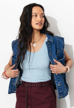 Femme portant un débardeur bleu clair, une veste en jean bleue sans manches, un pantalon bordeaux avec ceinture en corde, un collier chaîne en or avec pendentif en forme de fleur.