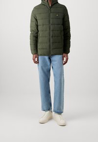 Grüne Daunenjacke mit Kapuze, ausgestattet mit einem Reißverschluss und horizontalem Steppmuster. Kombiniert mit hellblauen Jeans und weißen Turnschuhen.