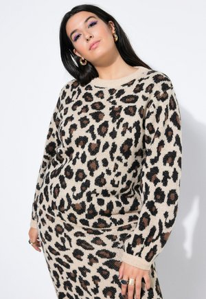 Pitkähiuksinen tummahiuksinen nainen käyttää kultaisia renkaankorvakoruja ja beigeä leopardi-kuvioista neulepusero- ja hameasua, poseeraa valkoista taustaa vasten.