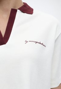 Άσπρο πόλο πουκάμισο με υφασμάτινη υφή και μπορντό γιακά και περιθώρια, με καλλιγραφική κέντηση στο στήθος.