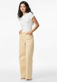 Pantalons beige à jambe large avec une taille haute, des poches avant et une fermeture à bouton en argent, associés à un T-shirt blanc ajusté.
