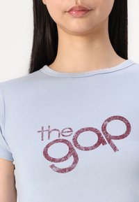 Camiseta ajustada de color azul claro en tejido suave, con un gráfico en color burdeos que dice "the gap" en el pecho, en un estilo de fuente desgastada.