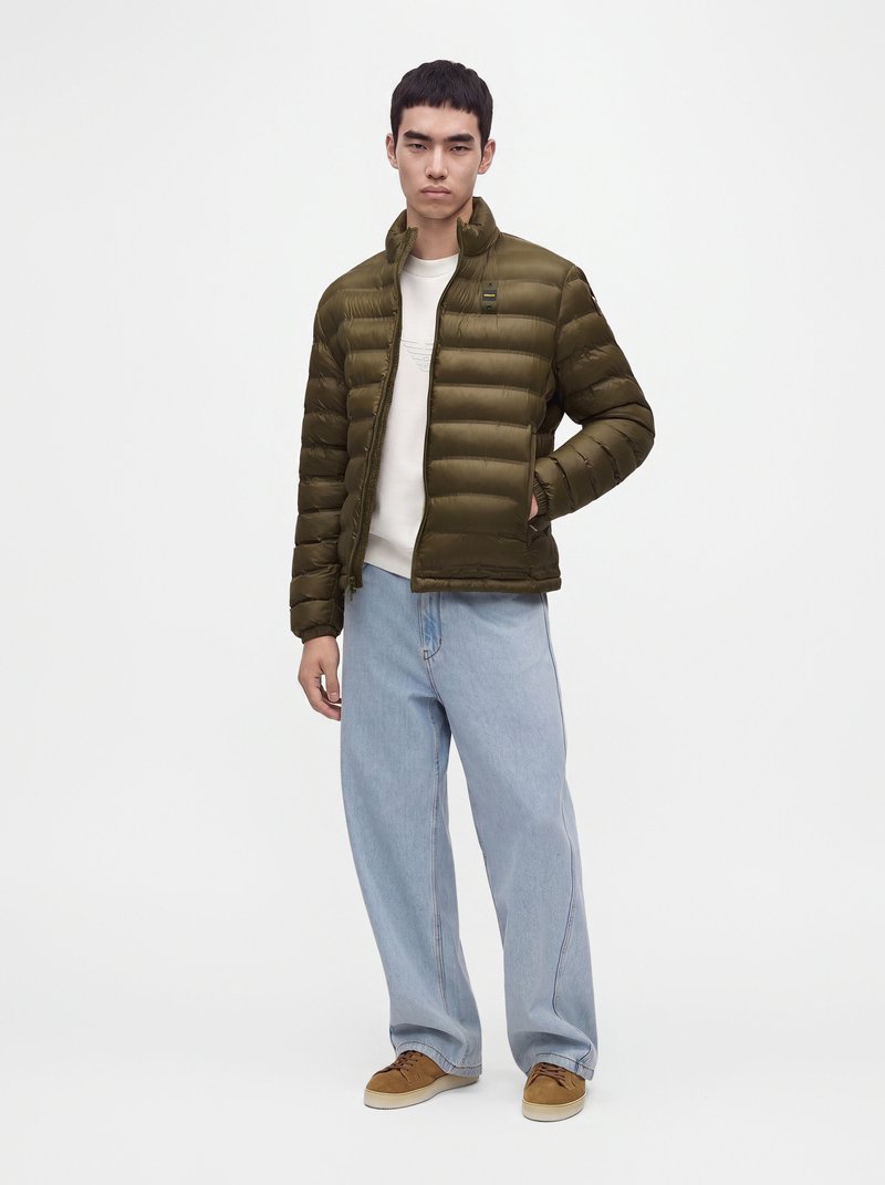 Homme portant une doudoune vert olive sur une chemise blanche, un jean large bleu clair et des baskets marron, debout devant un fond uni.