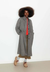 Un long manteau à carreaux gris et noir, avec un col arrondi, des poches avant et une fermeture à boutons. Assorti à un pull rouge, une jupe marron et des chaussures beiges.