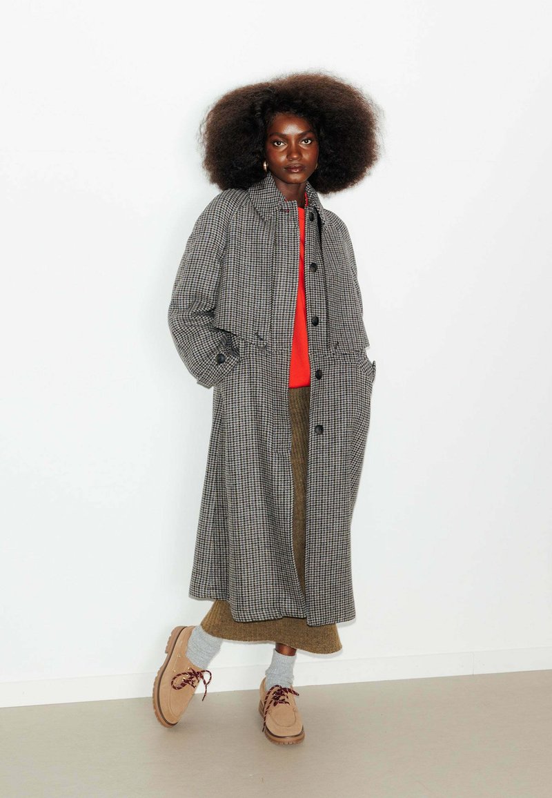 Un long manteau à carreaux gris et noir, avec un col arrondi, des poches avant et une fermeture à boutons. Assorti à un pull rouge, une jupe marron et des chaussures beiges.