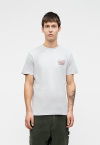 Γκρι βαμβακερό μπλουζάκι με κοντά μανίκια, με μια πολύχρωμη γραφιστική «Santa Cruz» στο αριστερό στήθος. Μοντέλο φορώντας πράσινα βελούδινα παντελόνια.