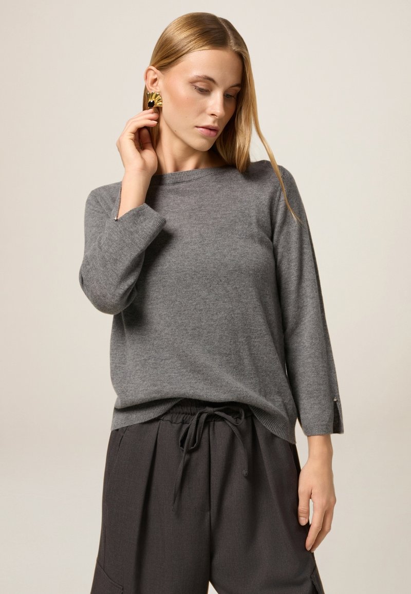 Grauer Strickpullover mit lockerer Passform, Rundhalsausschnitt und Dreiviertelärmeln. Mit dezenten Details an den Ärmelsäumen. Kombiniert mit dunklen Hosen.