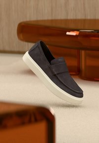 Σκούρο γκρι σουέντ παπούτσι χωρίς κορδόνια με λευκή παχιά σόλα, εμφανιζόμενο στον αέρα πάνω από ανοιχτό χαλί με έπιπλα κεχριμπαρένιου διαφανούς χρώματος στο φόντο.