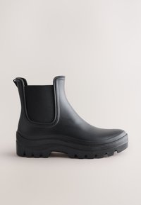Black ankle rain boot with thick treaded sole and elastic side panels on light background.