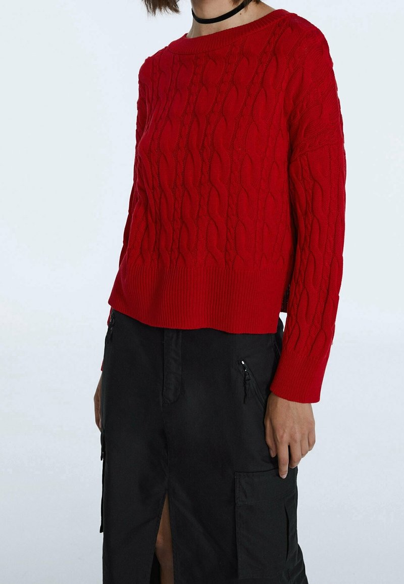 Pullover rouge en tricot torsadé avec un col ras du cou côtelé et des manches longues, associé à une jupe noire avec des zips et une fente latérale.