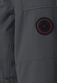 Chaqueta deportiva de tela gris con un parche redondo de logo negro y rojo, costuras visibles y una superficie texturizada.