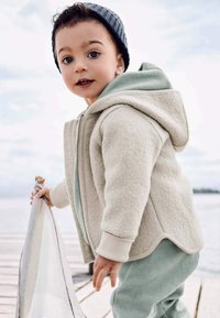 Kleinkind in beiger Jacke und grauer Mütze, das auf einem Holzsteg nahe am Wasser steht und einen hellen Stoff oder eine Decke hält.
