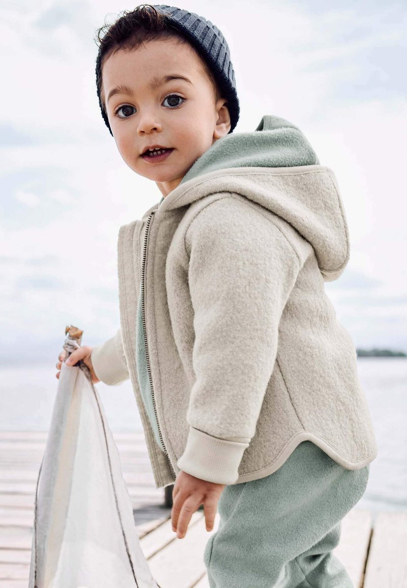 Kleinkind in beiger Jacke und grauer Mütze, das auf einem Holzsteg nahe am Wasser steht und einen hellen Stoff oder eine Decke hält.