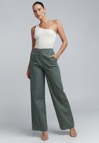 Witte geribbelde bodysuit met één schouder. Groene wijde broek met zijzakken en verticale naden, gecombineerd met transparante hakken.