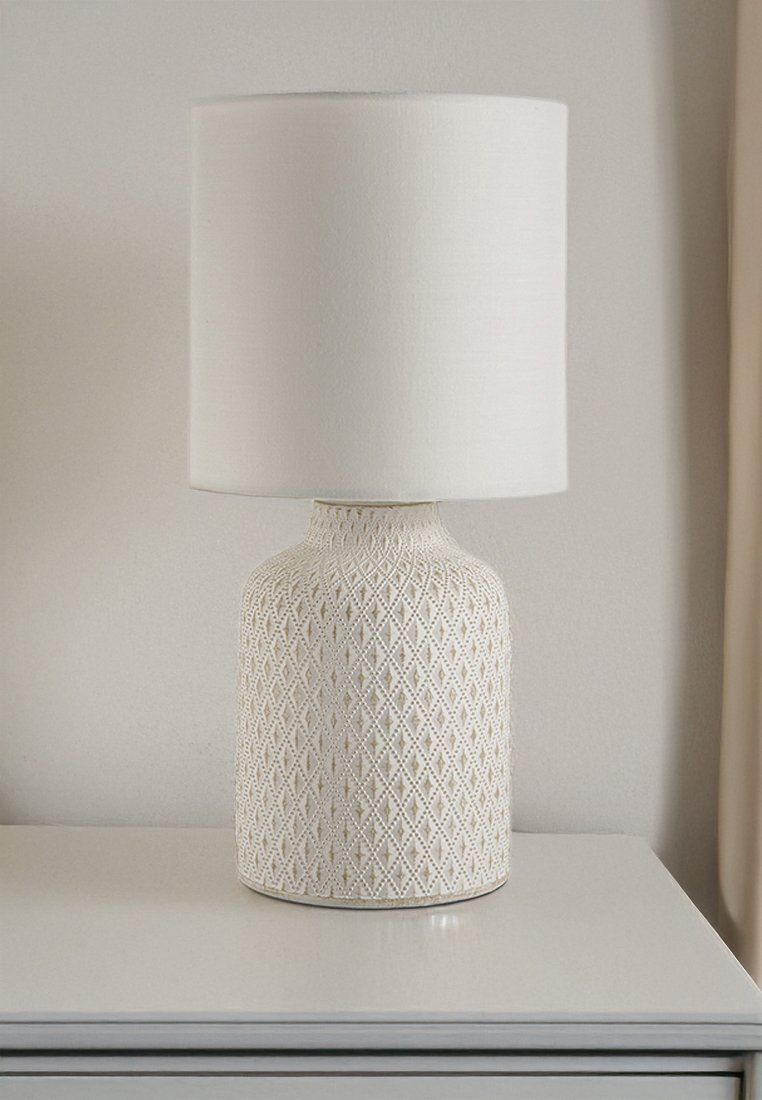 Bílá keramická stolní lampa se základnou se vzorem diamantů a jednoduchým válcovým látkovým stínidlem na bílém povrchu před světle zbarvenou zdí.