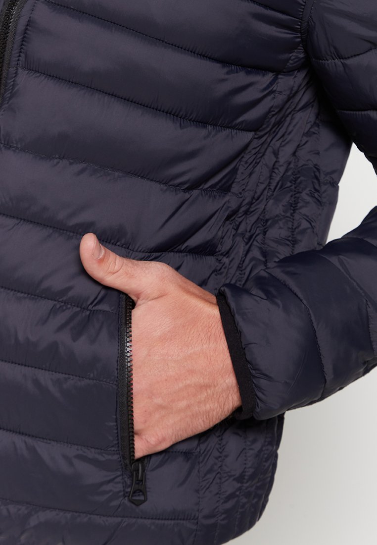 Marine pufferjack met een gequilte textuur, ritszak en zwarte accenten op de mouwen. De materialen lijken lichtgewicht en geïsoleerd te zijn.