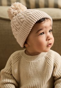 Beige gestrickte Mütze mit Bommel oben, mit einem strukturierten Zopfmuster; kombiniert mit einem gemütlichen beigen Strickpullover.