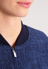 Marineblaue, strukturierte Jacke mit durchgehendem Reißverschluss, geripptem Kragen und glattem Reißverschlussgriff. Nahaufnahme des Halsbereichs.